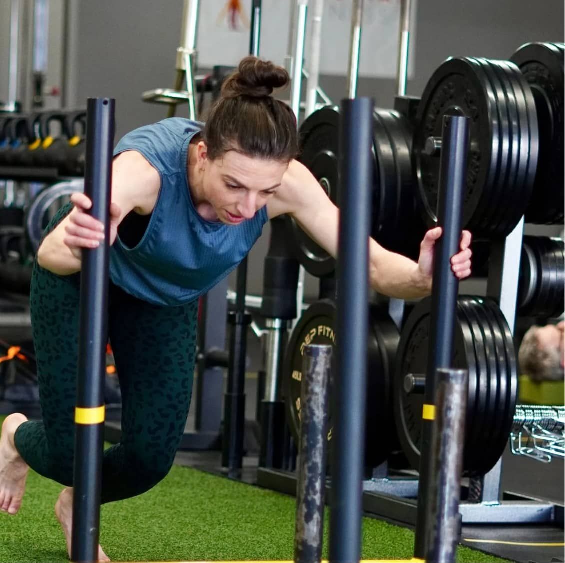 A woman pushing a sled on a gym's artificial turf, wearing a blue tank top and green leggings. She appears focused and determined. Fitness equipment and weights are in the background.