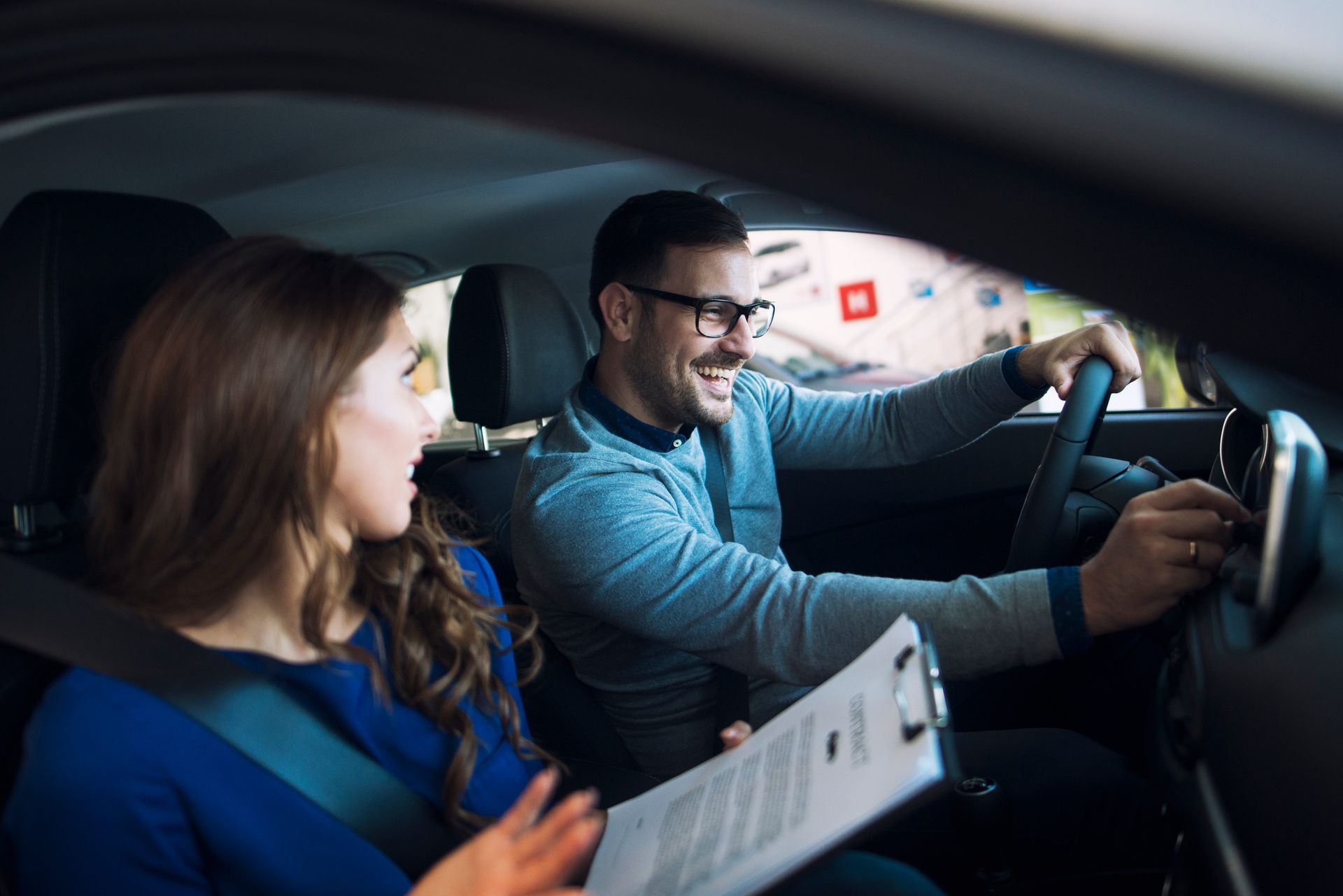 A smiling driver in a blue shirt holds up a driver's license, giving a thumbs up alongside an instructor in a car.