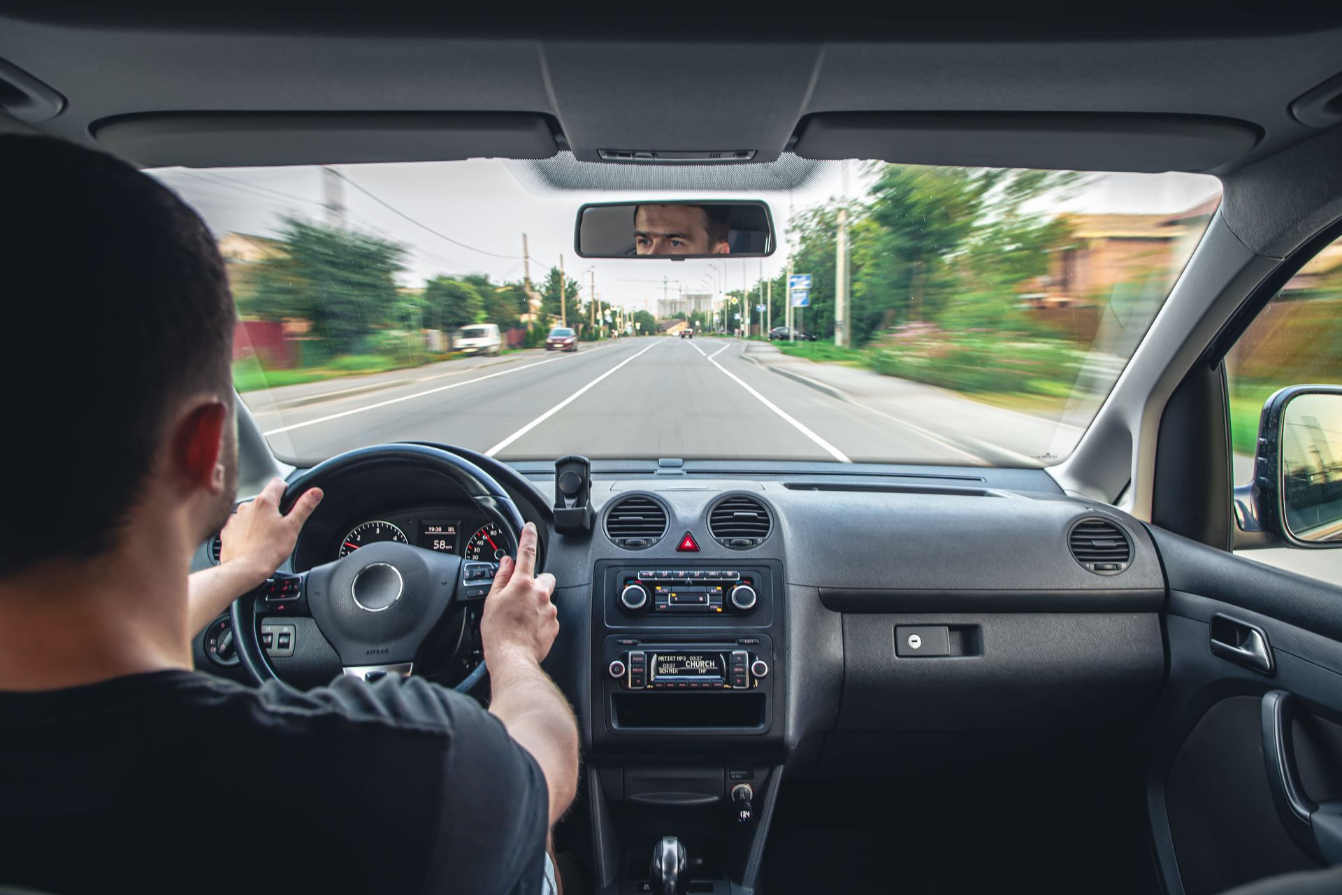 A driving instructor points at the road while a student steers a car during a lesson, viewed from the backseat.