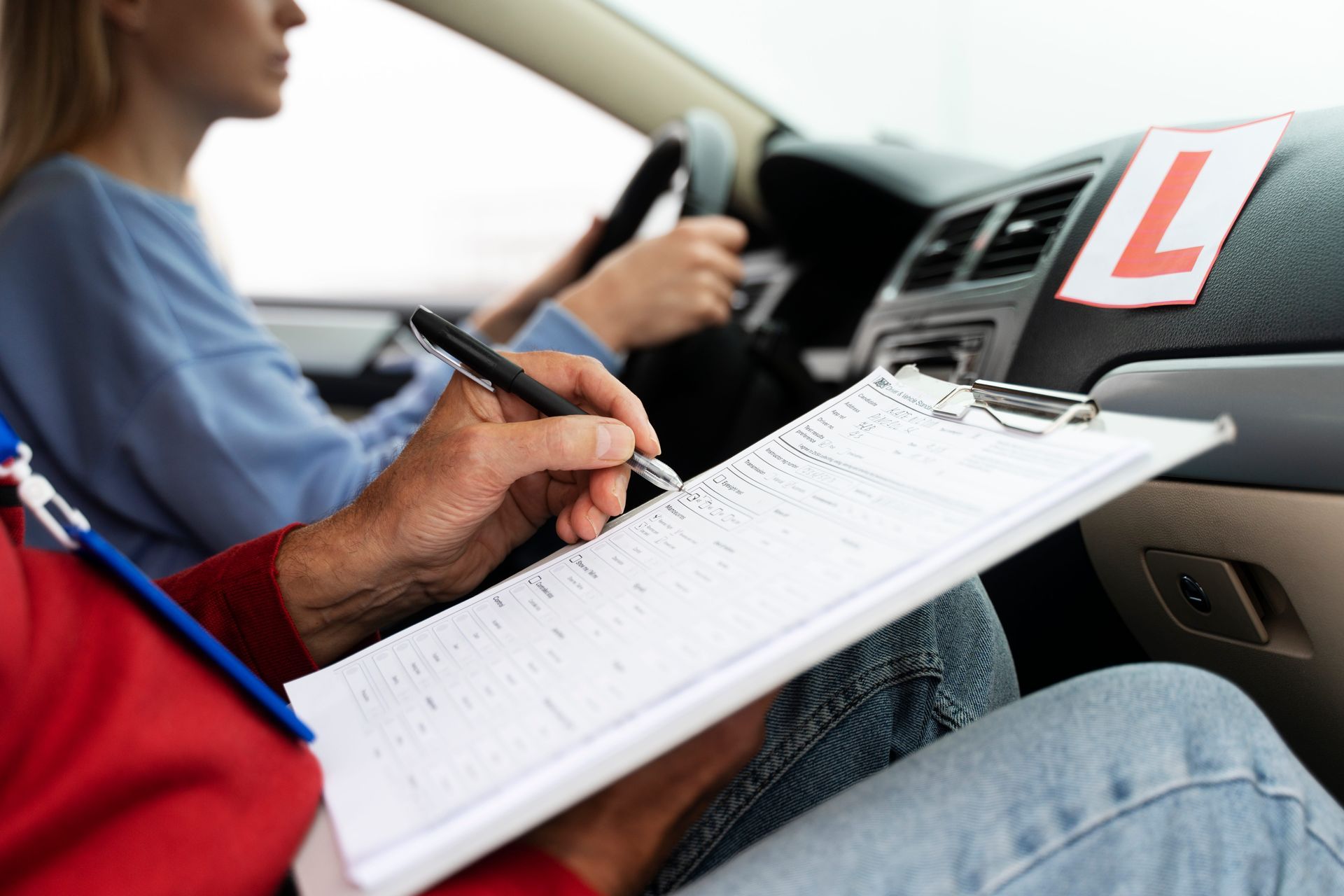 A smiling driver in a blue shirt holds up a driver's license, giving a thumbs up alongside an instructor in a car.