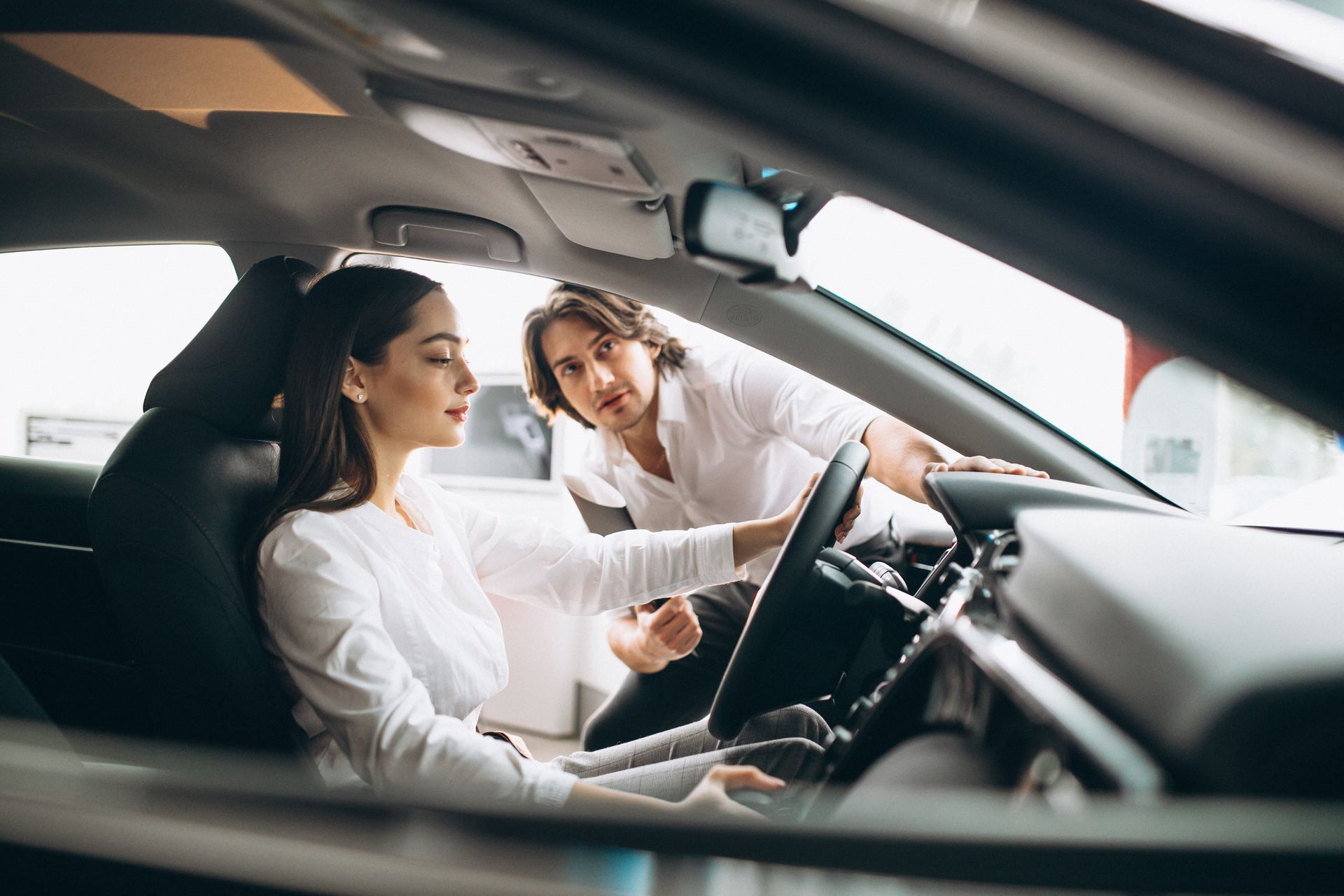 A driving instructor holding a blue clipboard gives directions to a person driving a car.