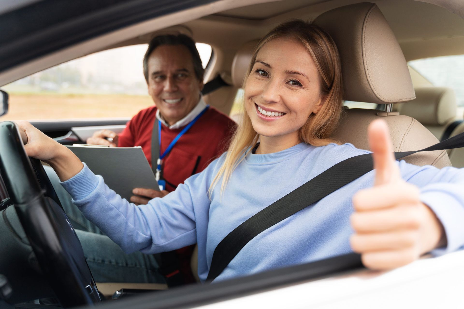 Two people laughing together while seated in a car, with the driver in a striped shirt and the passenger wearing a hat.
