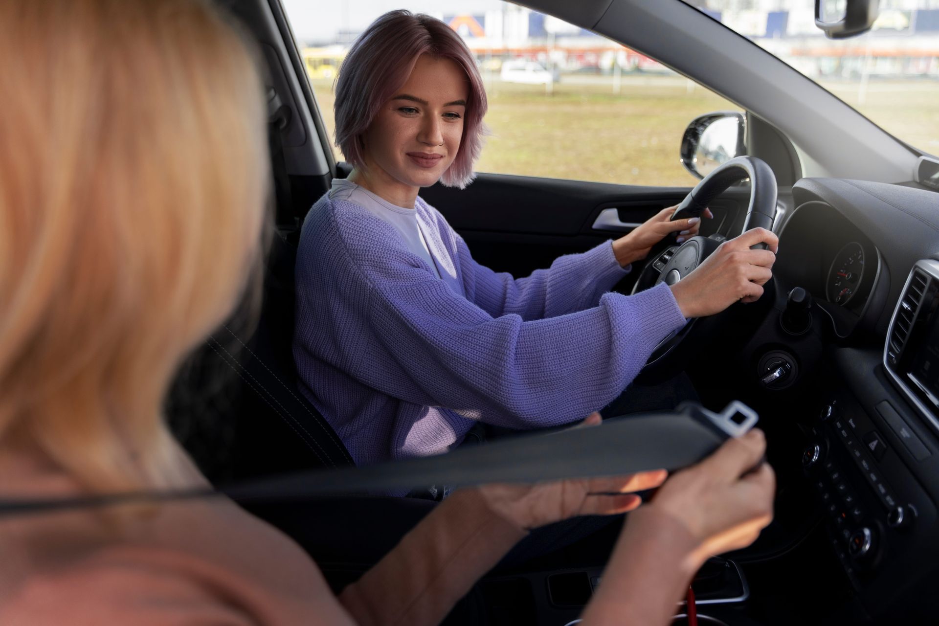 A smiling driver in a blue shirt holds up a driver's license, giving a thumbs up alongside an instructor in a car.