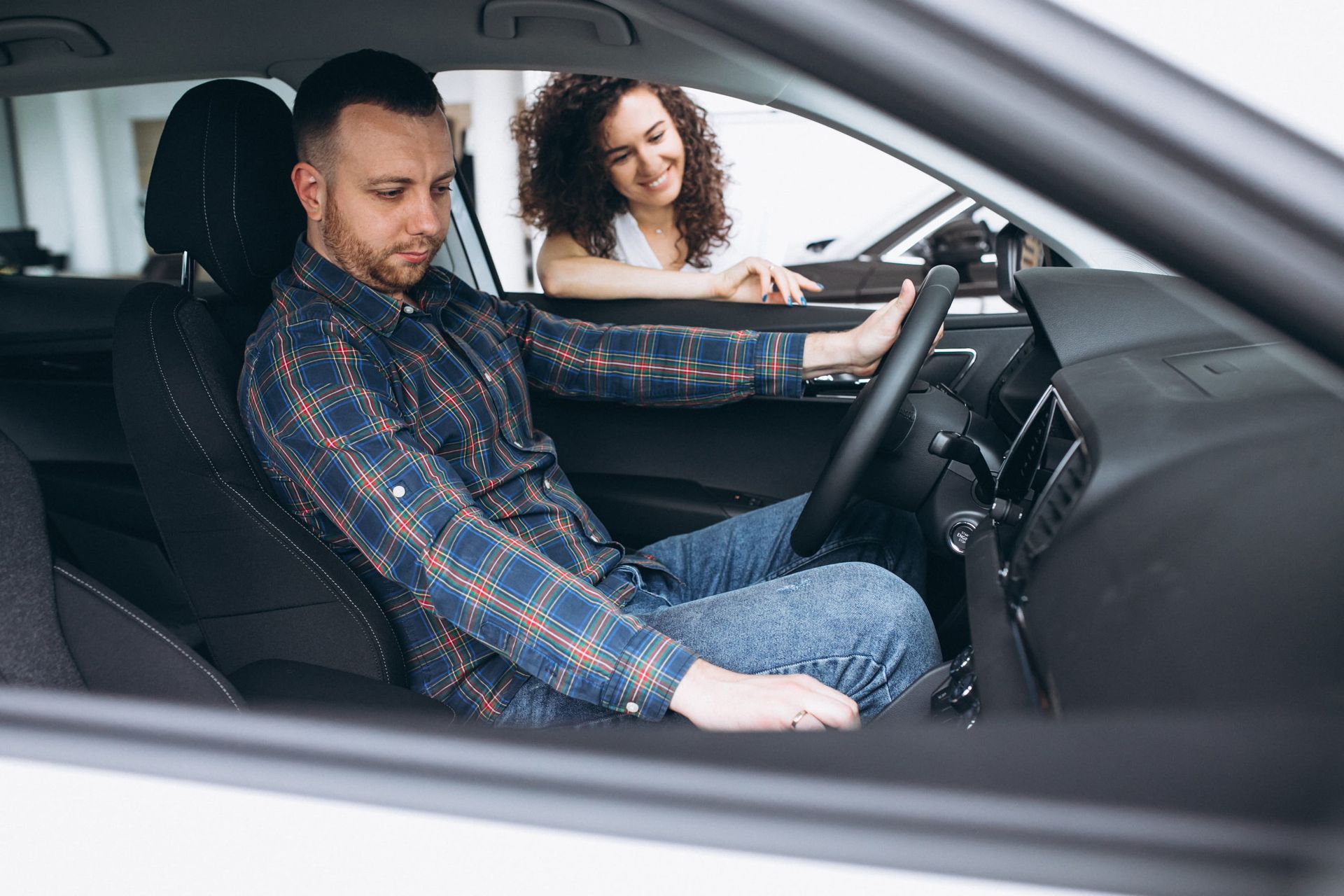A driving instructor holding a blue clipboard gives directions to a person driving a car.