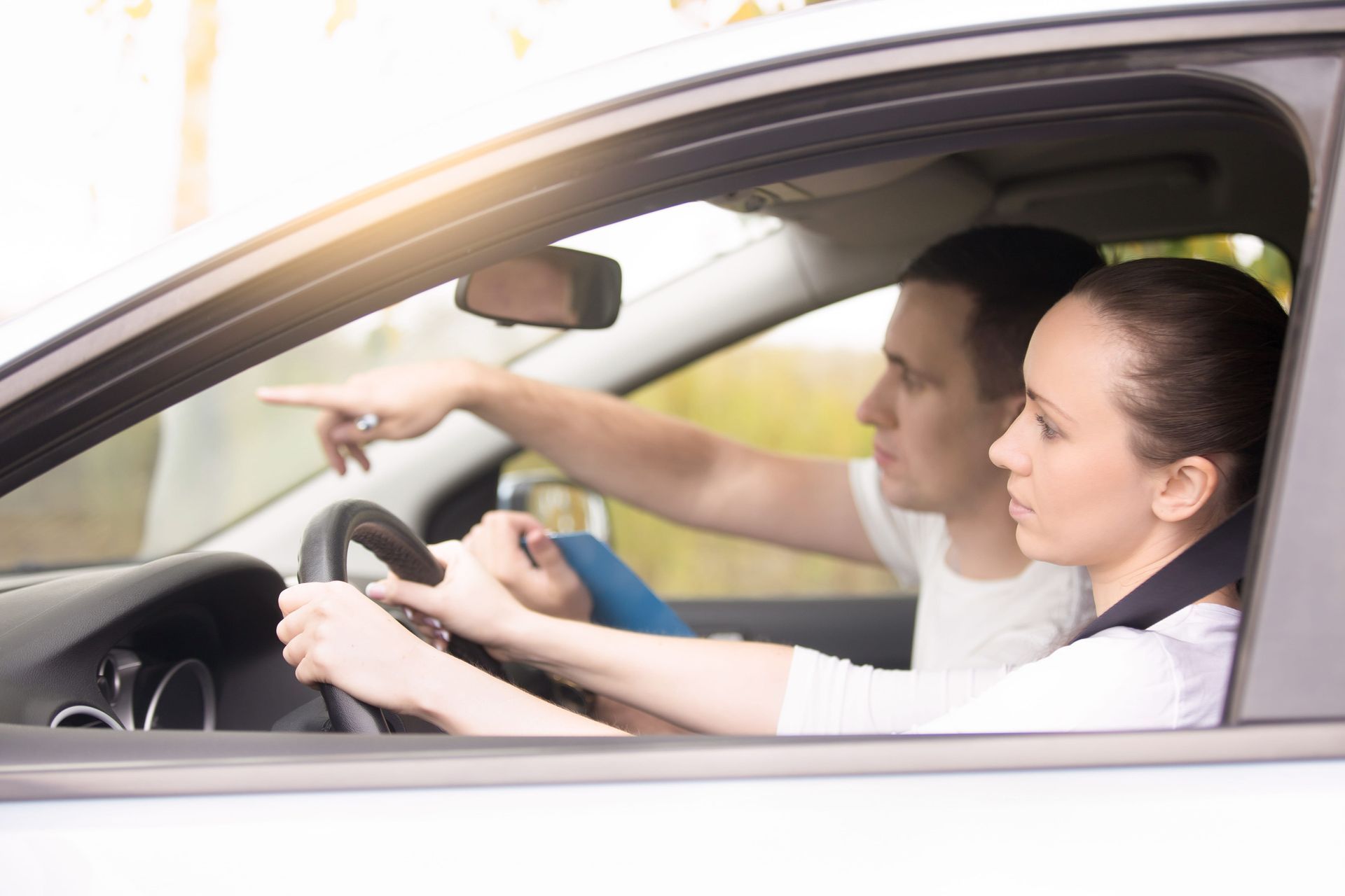 A driving instructor points at the road while a student steers a car during a lesson, viewed from the backseat.