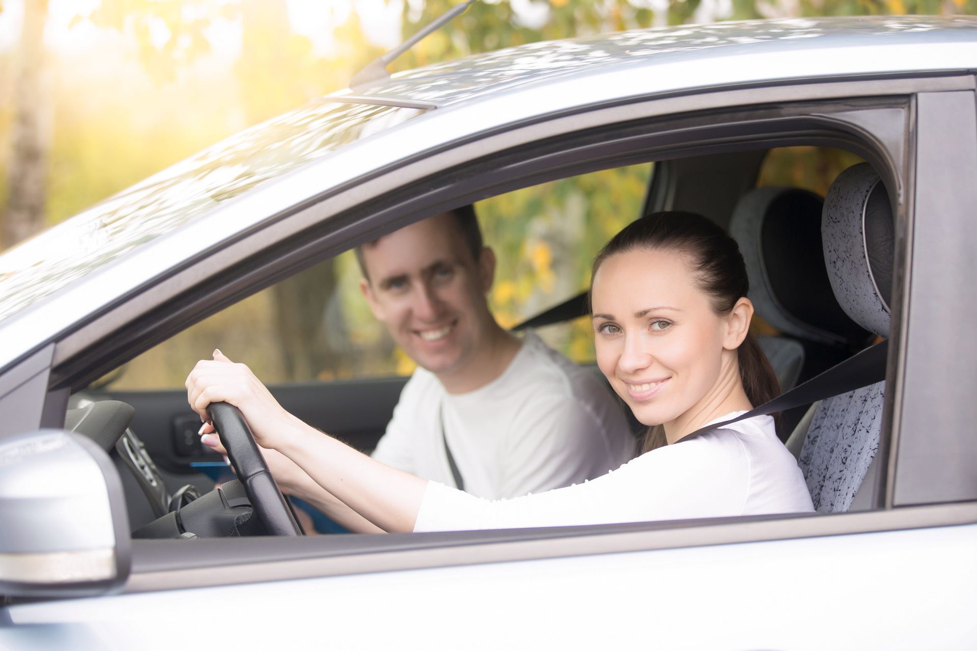 A smiling driver in a blue shirt holds up a driver's license, giving a thumbs up alongside an instructor in a car.