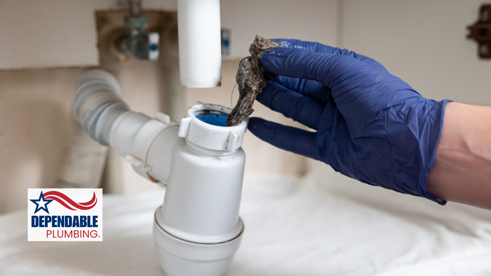 Gloved hand removing a clog from a white sink drain pipe, showing debris. The scene is inside, and a logo is visible.