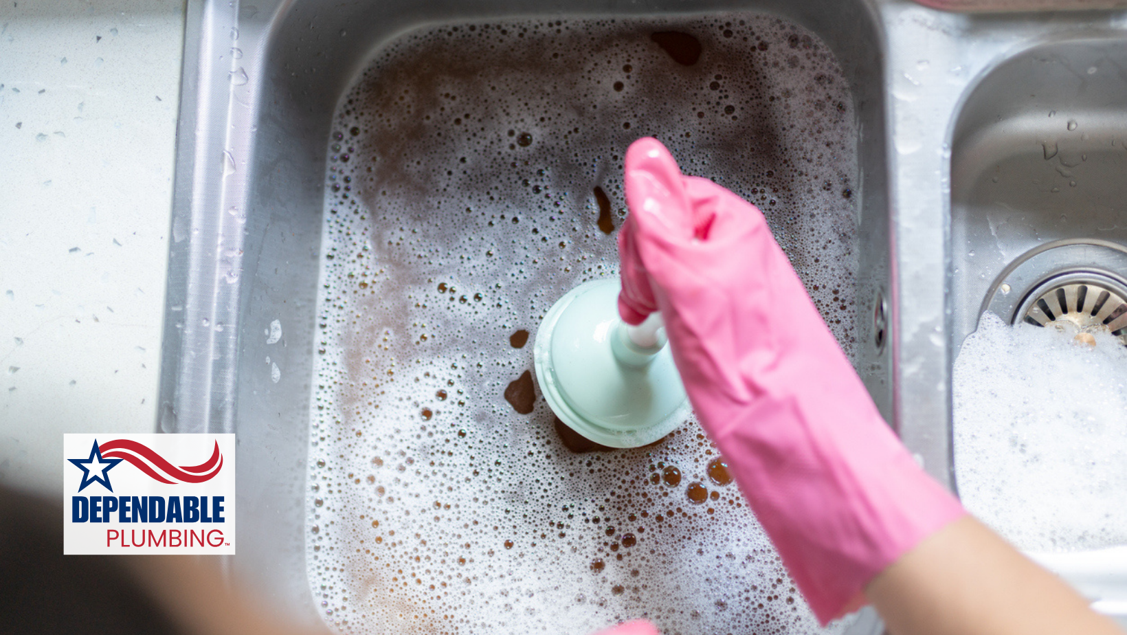 Person using a plunger in a soapy kitchen sink, wearing pink gloves.