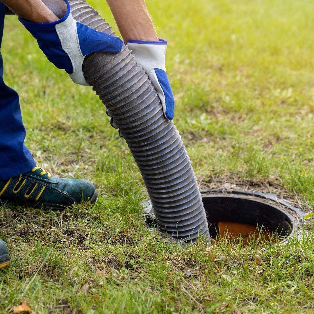 Person in blue gloves and overalls inserting a hose into a septic tank opening in a grassy yard.