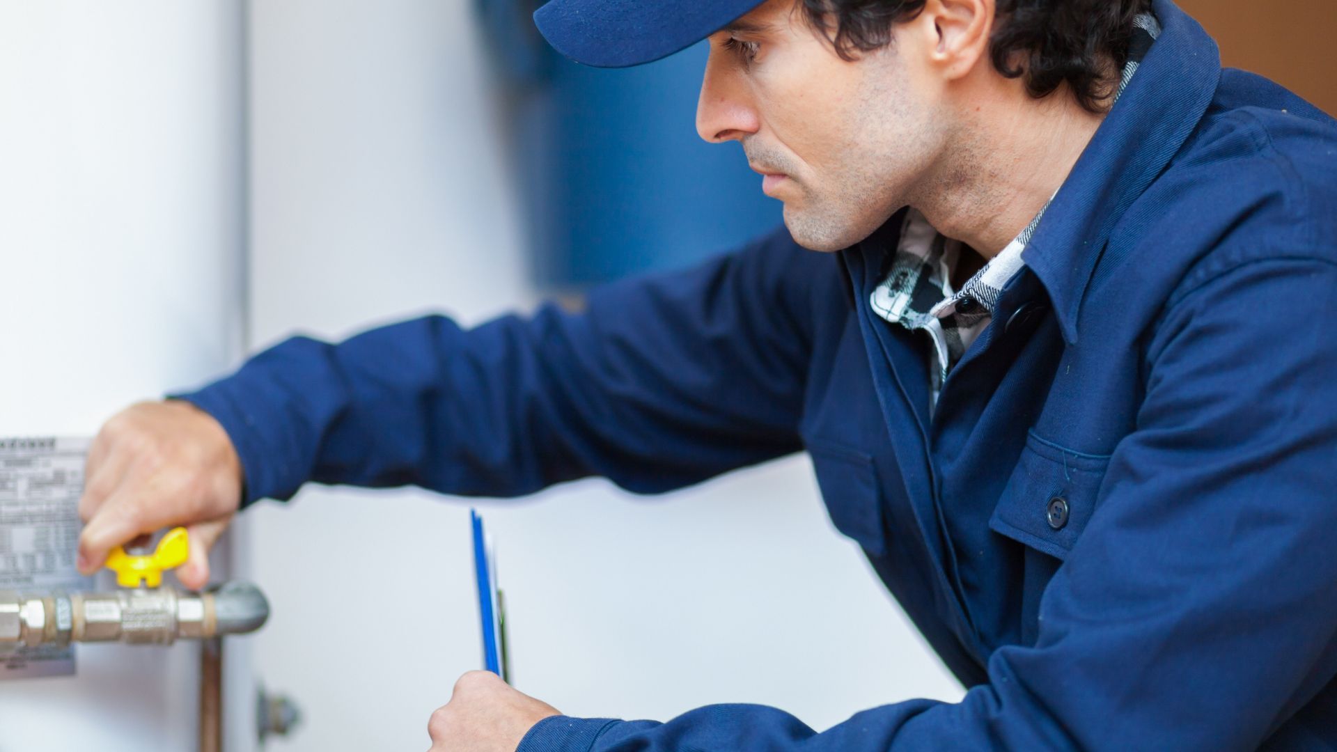 A man in a blue jumpsuit, adjusting plumbing with a wrench, indoors.