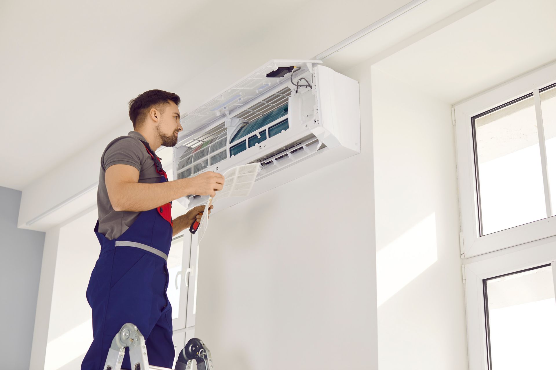 A man is standing on a ladder fixing an air conditioner.