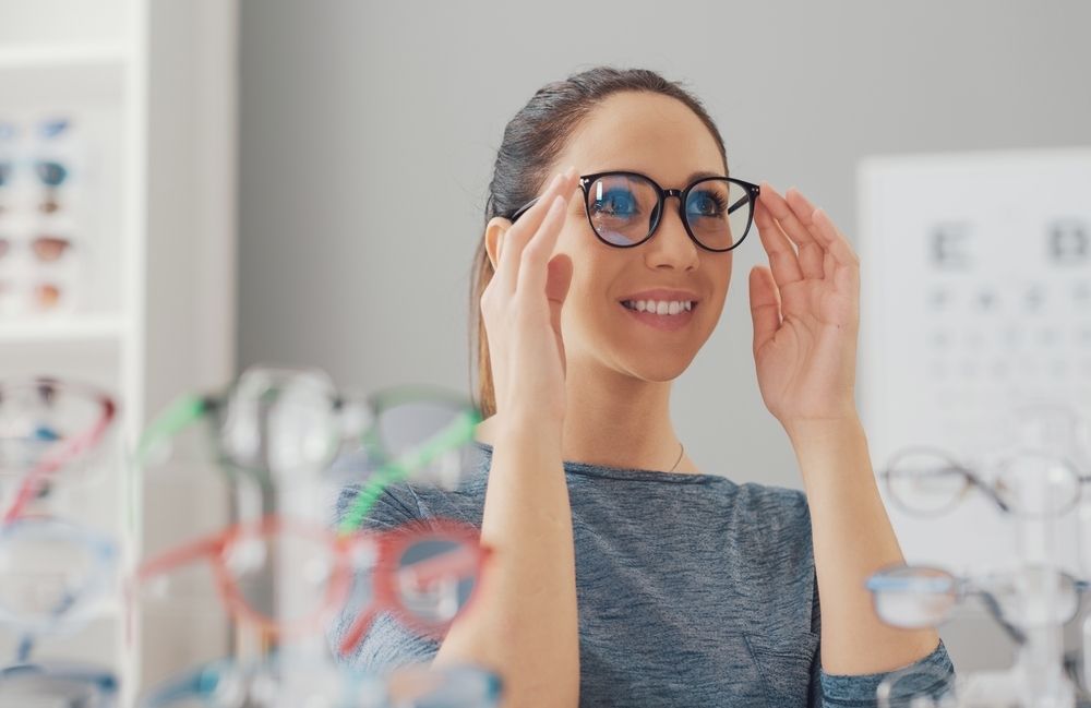 Woman smiles, trying on black glasses in an optician's office. Shelves of glasses in background   — Eyepower In Tahmoor, NSW