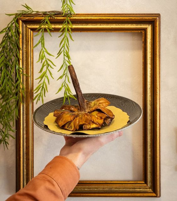 Mano che regge un piatto gourmet impreziosito da un'elegante ghirlanda, incorniciato in oro e sormontato da tralci di vite verdi.