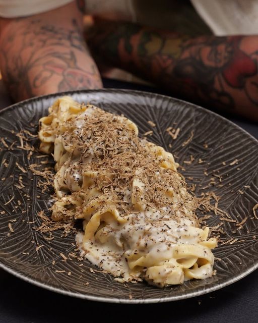 Pasta cremosa su un piatto scuro, guarnita con erbe aromatiche, davanti a braccia tatuate su un tavolo