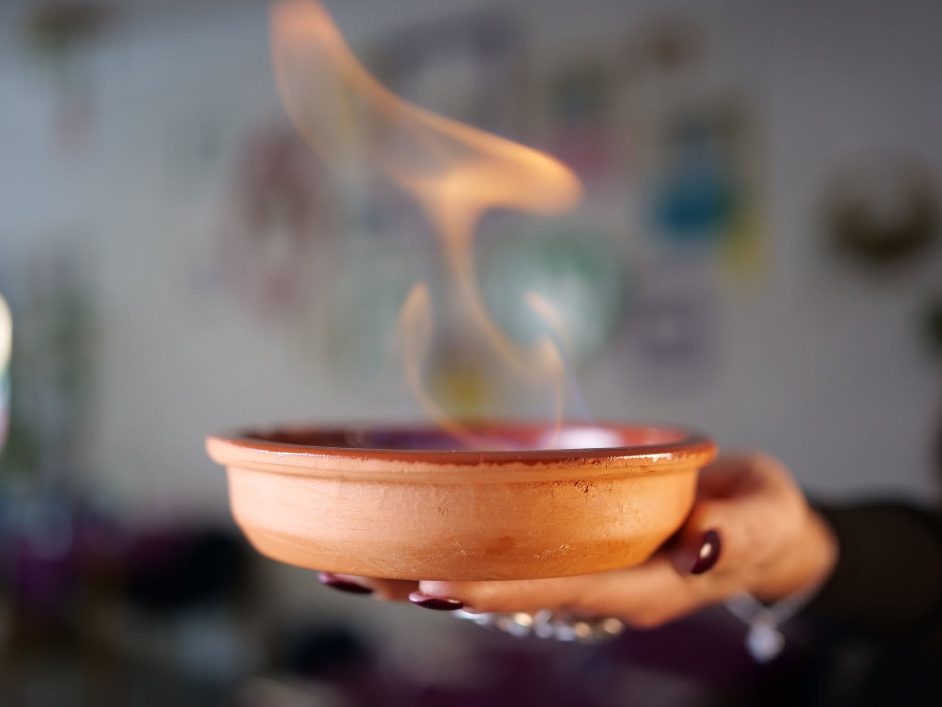 Una mano che regge una ciotola di terracotta da cui si sprigiona una fiamma accesa, in un ambiente interno.