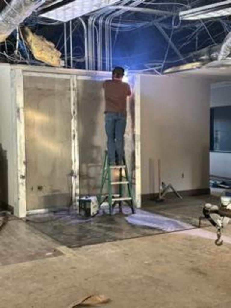 Person on a step ladder painting a wall in a room under construction, with exposed ceiling above.