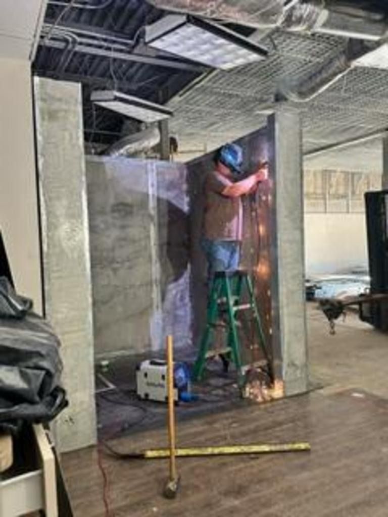 Worker on ladder welding inside a transparent barrier in an unfinished industrial space