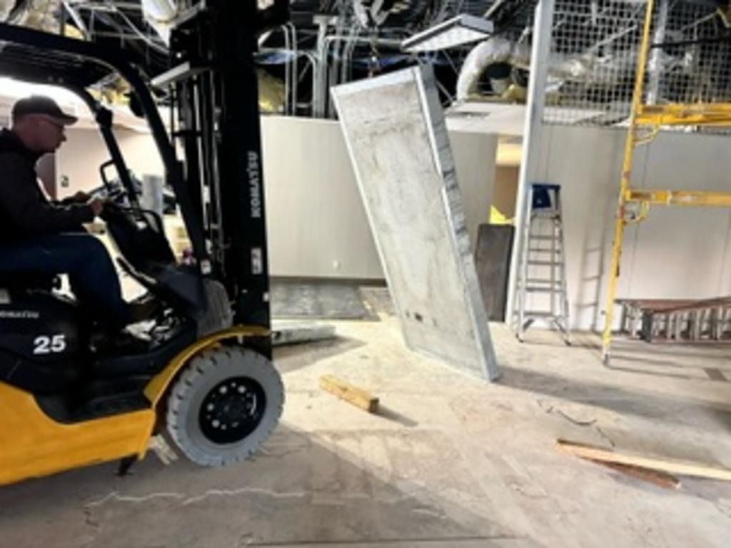 Forklift inside an industrial warehouse lifting a large metal panel near a ladder and shelving.