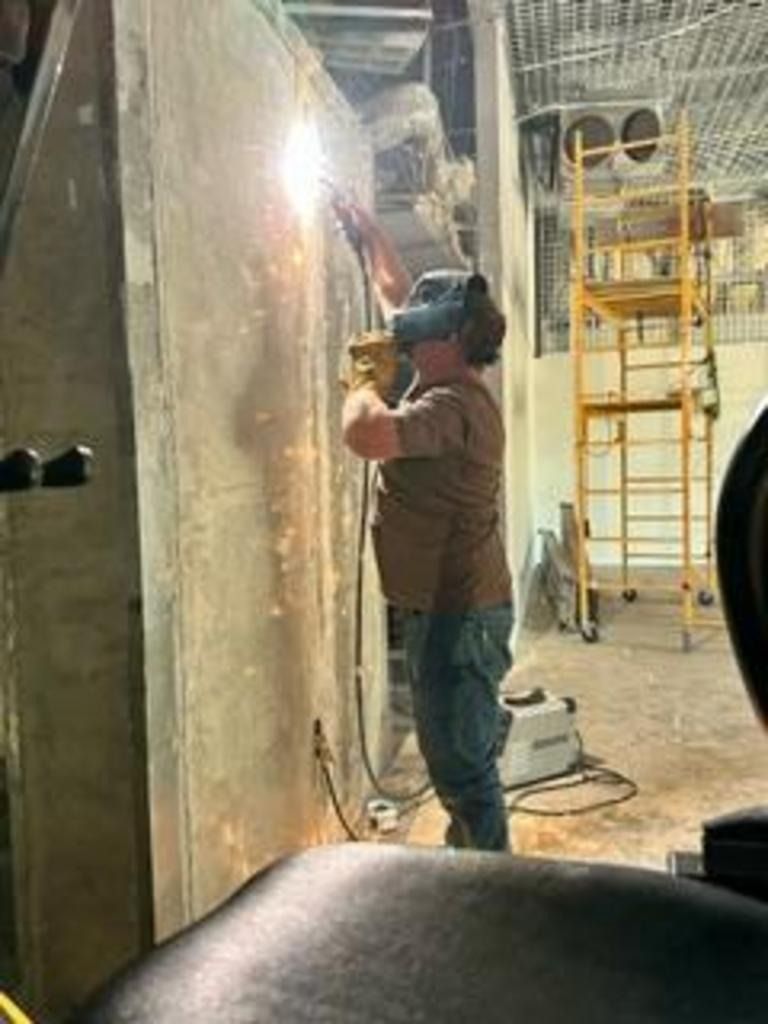 Person welding a large metal structure in a workshop, with sparks and industrial equipment nearby