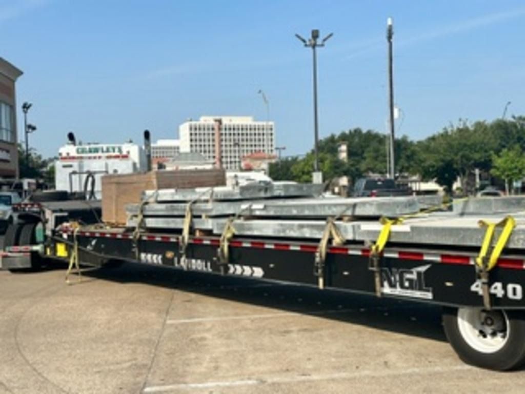 Black flatbed trailer loaded with stacked metal pipes in a parking lot under a clear sky