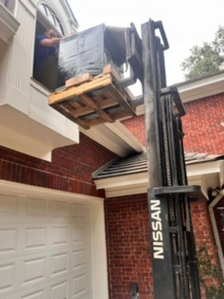 Forklift lifting a wrapped pallet to a second-story balcony beside a brick house