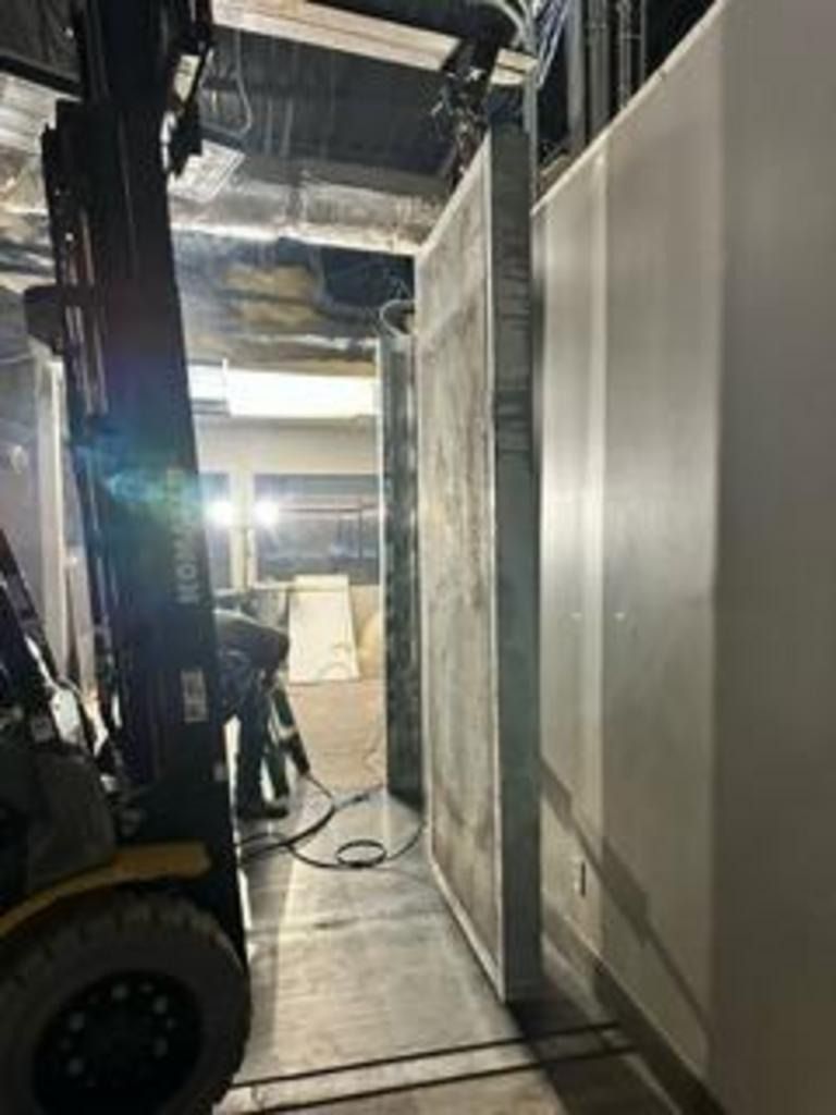 Forklift in a narrow industrial hallway beside a large wall panel and hanging ceiling materials.