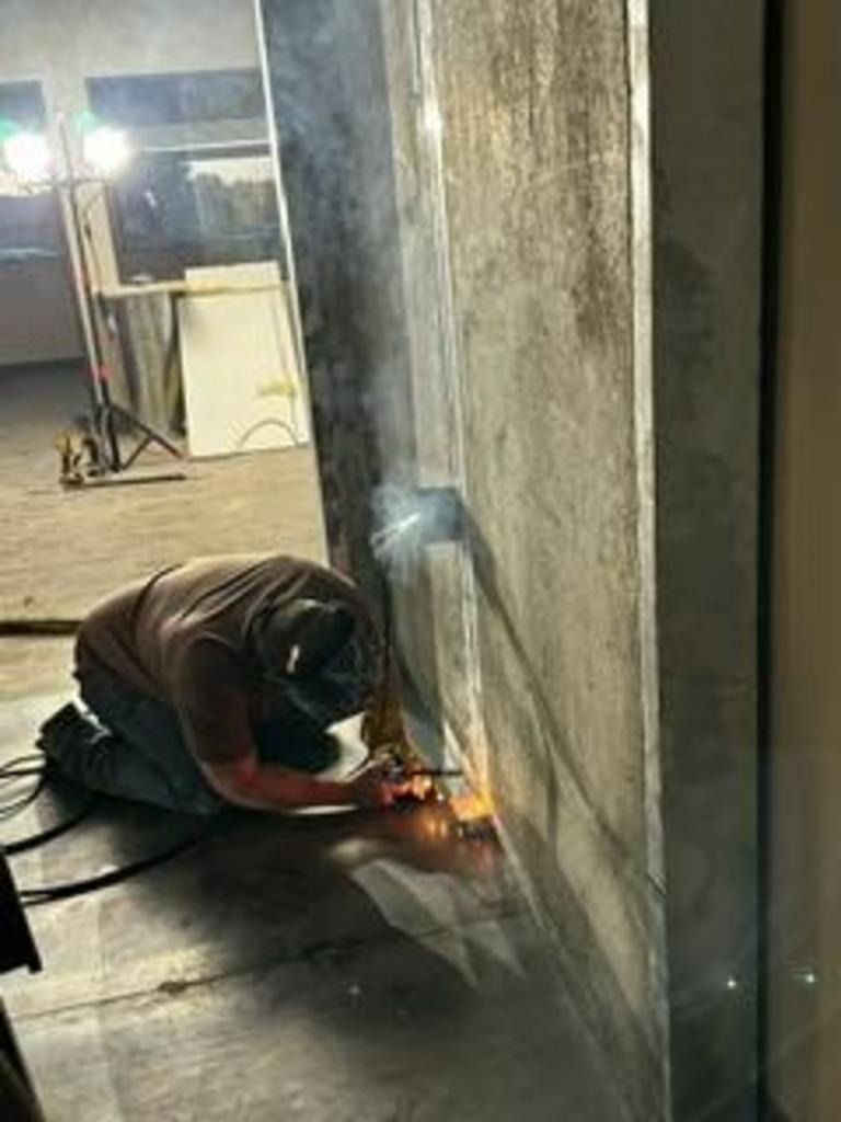 Worker welding along a concrete wall in a dim industrial room, sparks visible.