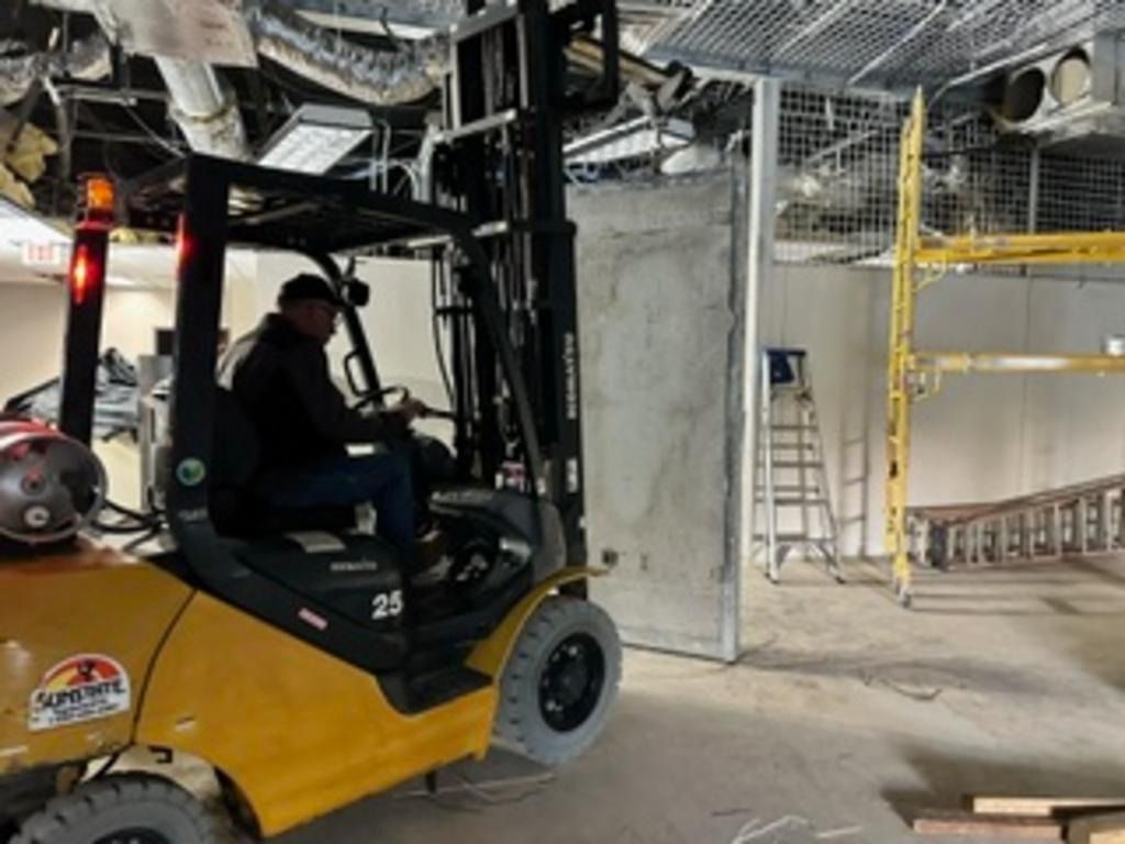 Forklift operator inside a construction site with exposed pipes, scaffolding, and a ladder.