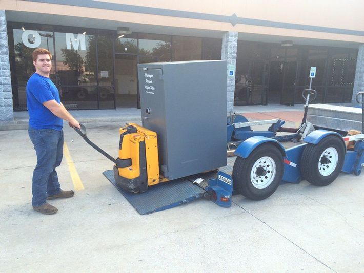 Man moving a yellow pallet jack with a large gray cabinet loaded on a trailer outside a building