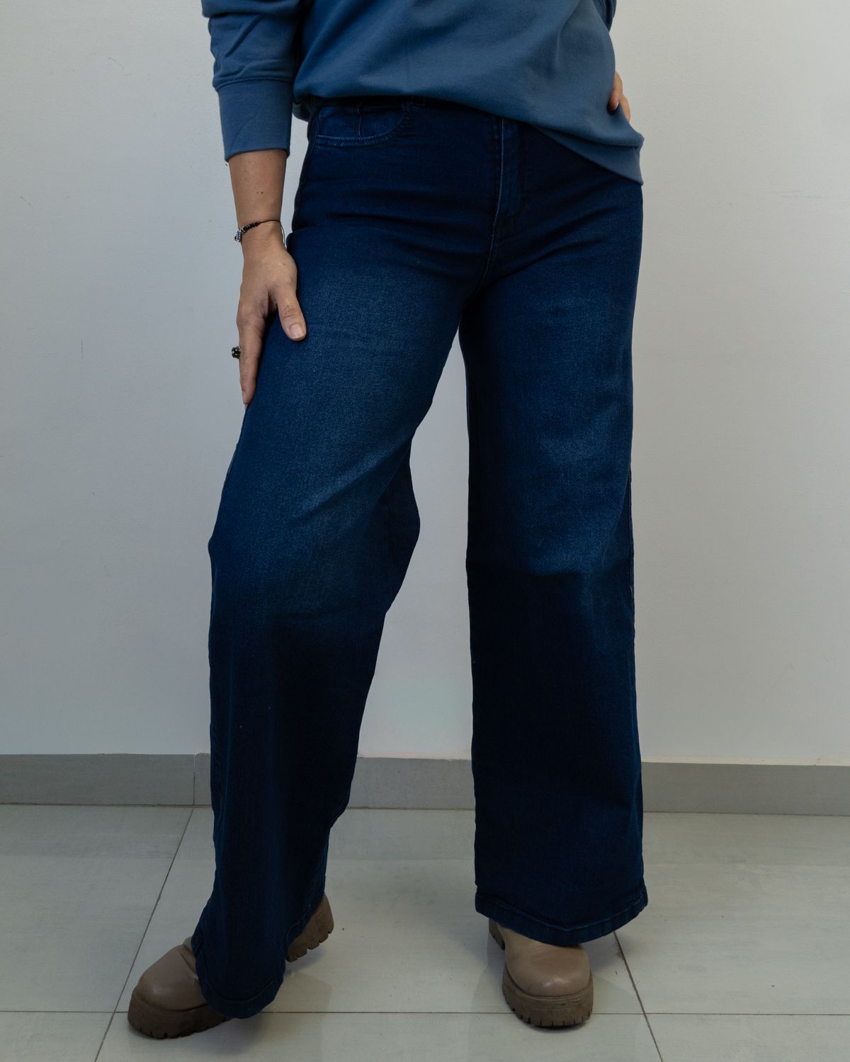 Persona que viste jeans azules y un suéter azul, parada frente a una pared blanca.
