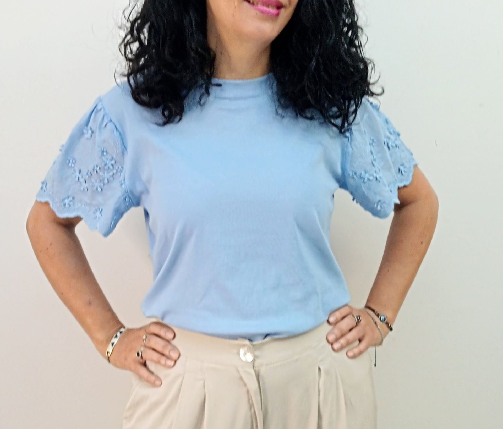 Mujer con un top azul claro con mangas de ojal y pantalones beige, con las manos en las caderas.