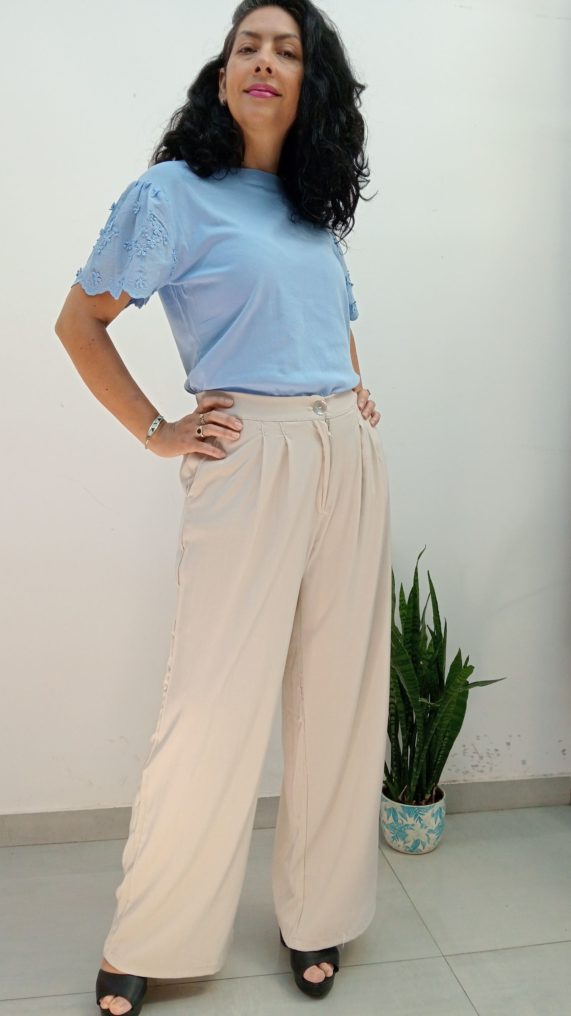 Mujer con un top azul claro con mangas de ojal y pantalones beige, con las manos en las caderas.