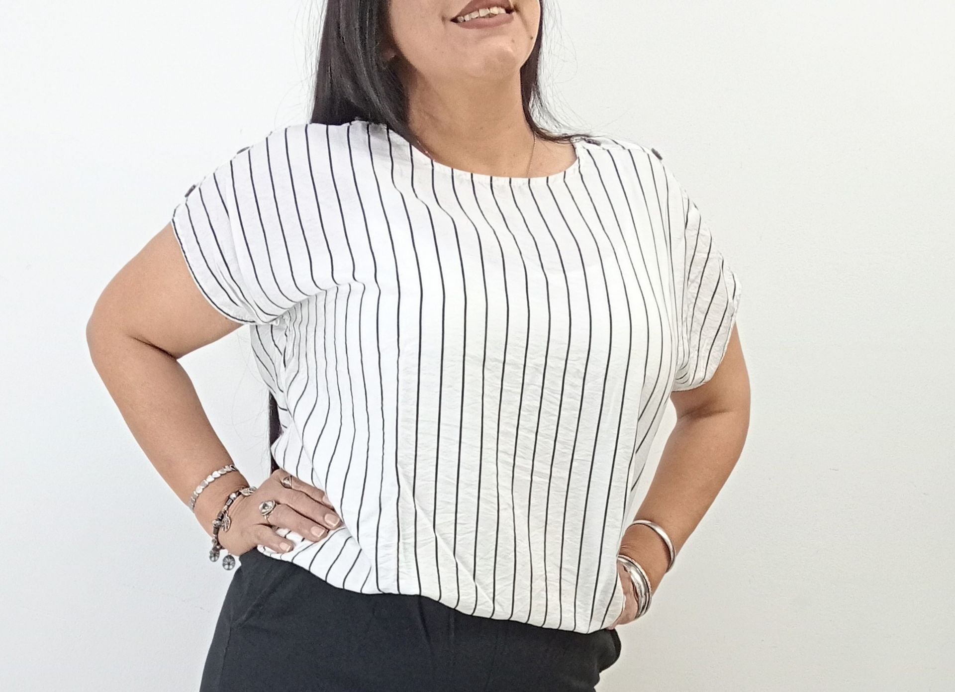 Mujer con camisa de rayas y pantalón negro, con las manos en las caderas, sonriendo, contra una pared blanca.
