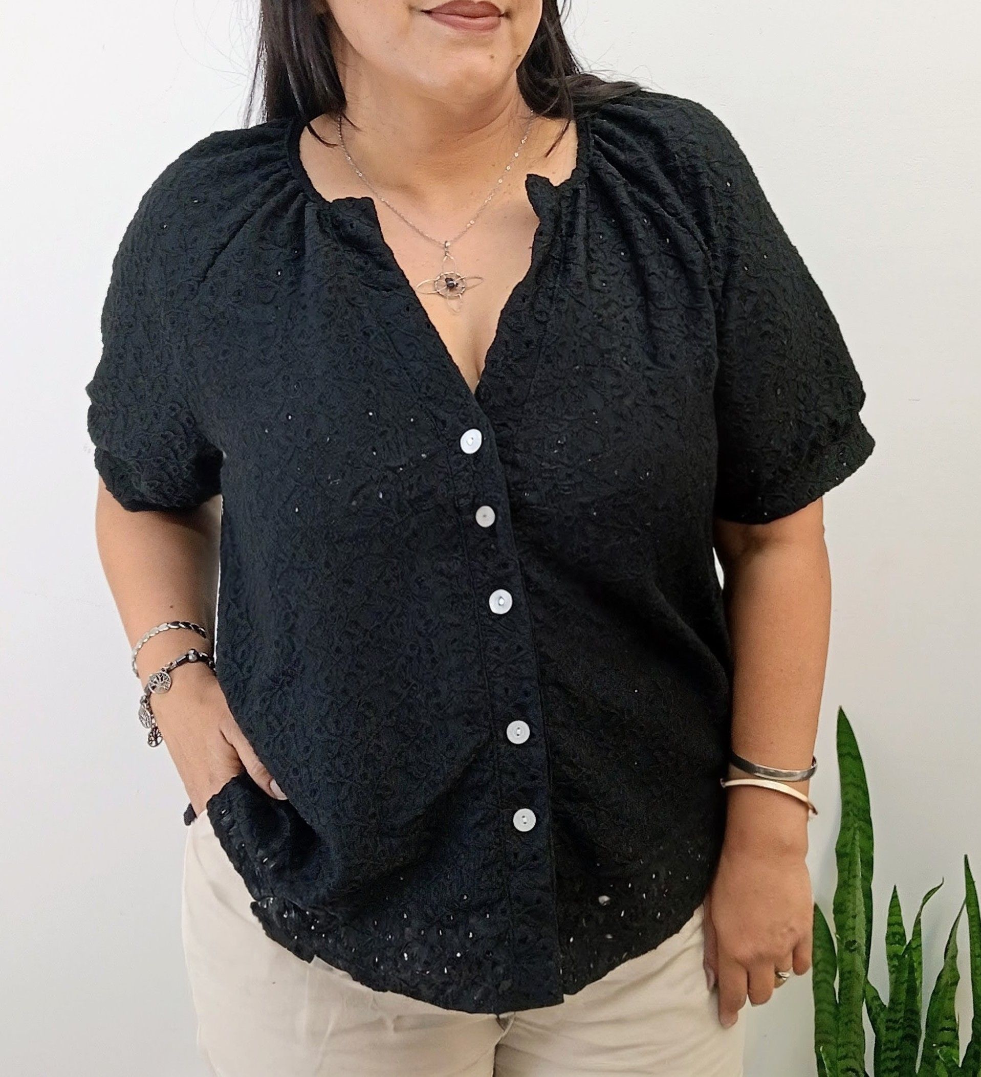 Mujer con camisa negra abotonada y pantalones beige, con joyas, de pie cerca de una planta.