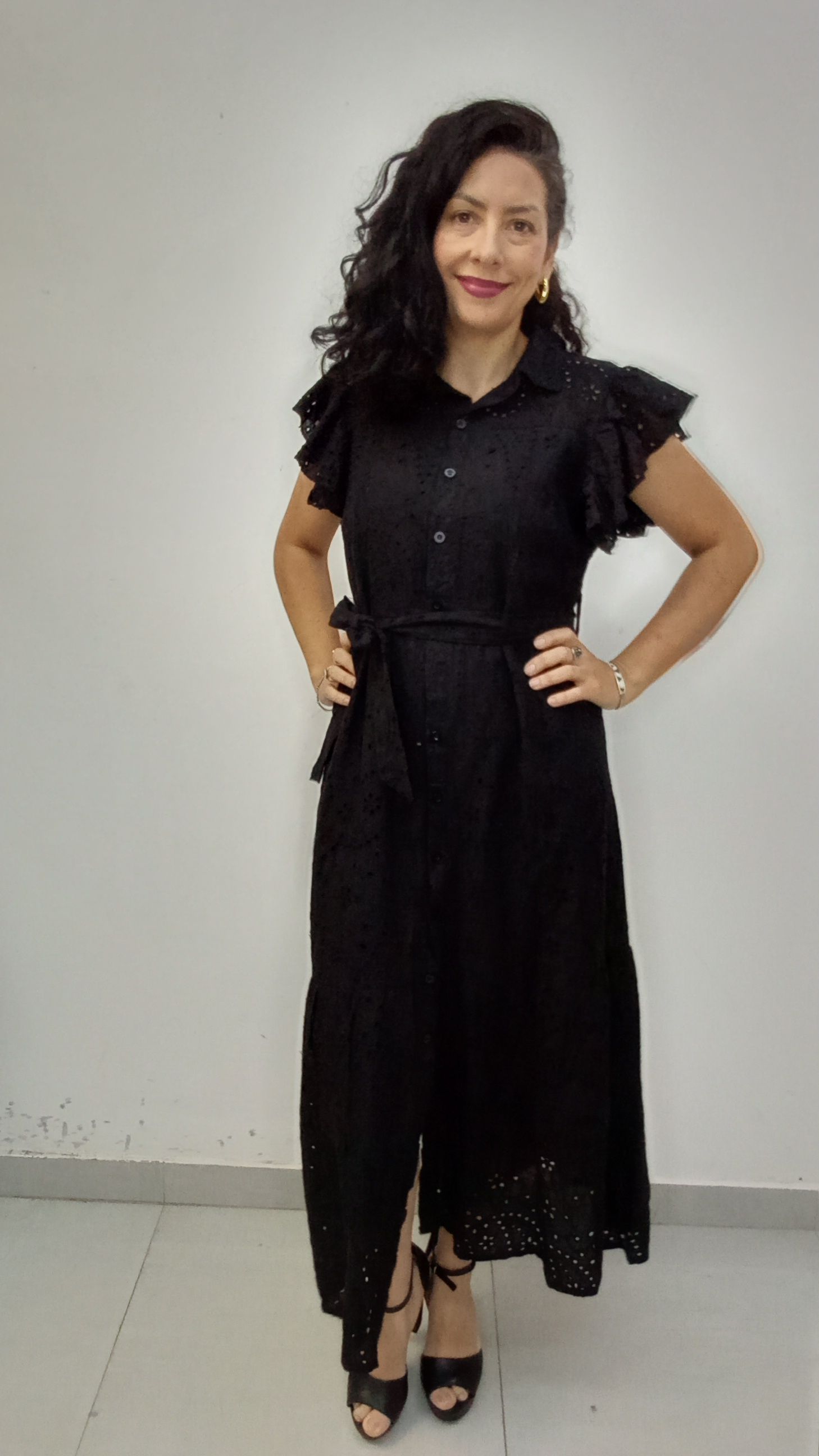 Mujer con vestido negro con detalles de ojales, manos en las caderas, sonriendo.