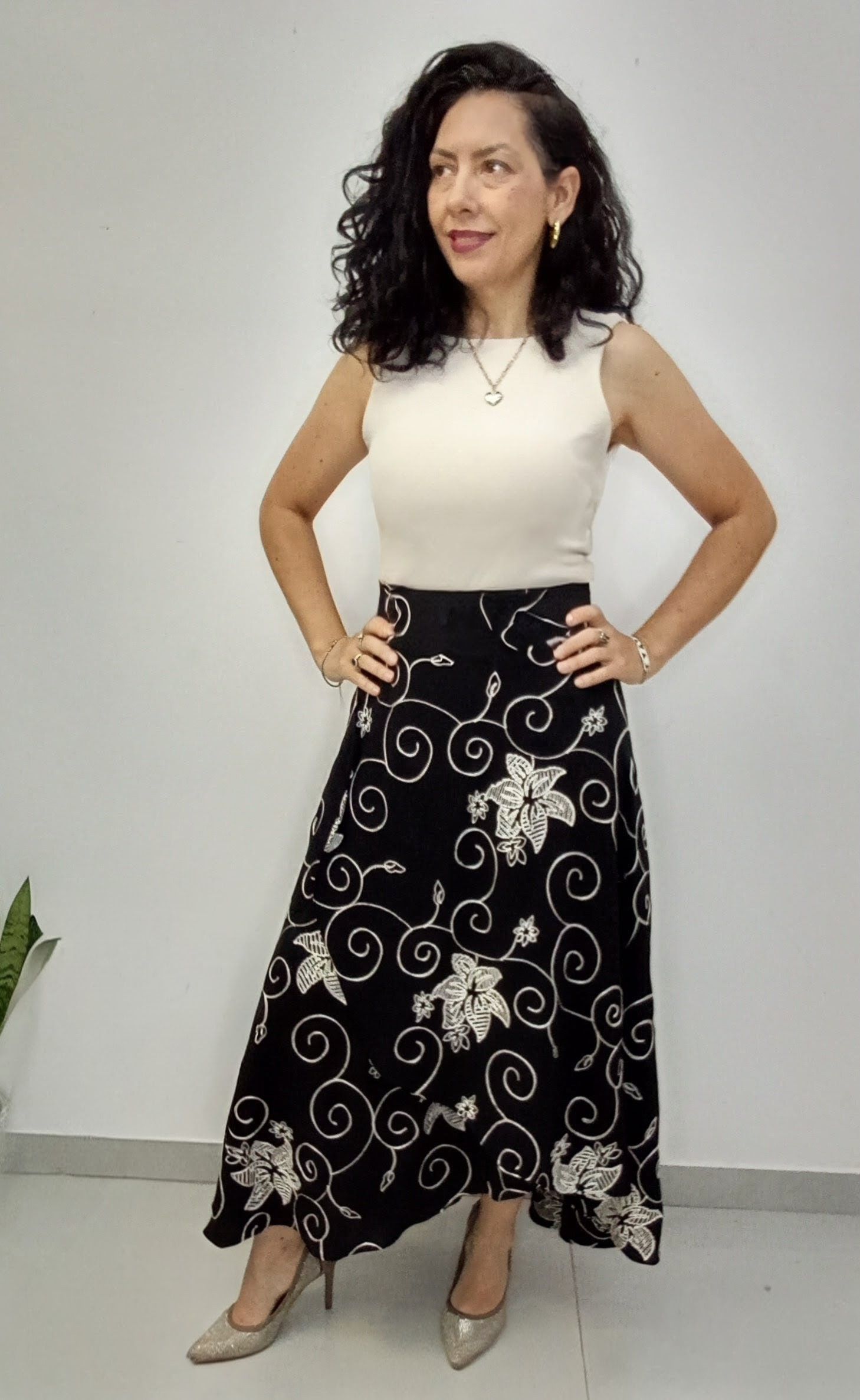 Mujer con top crema sin mangas y falda floral negra, manos en las caderas, sonriendo, usando tacones.