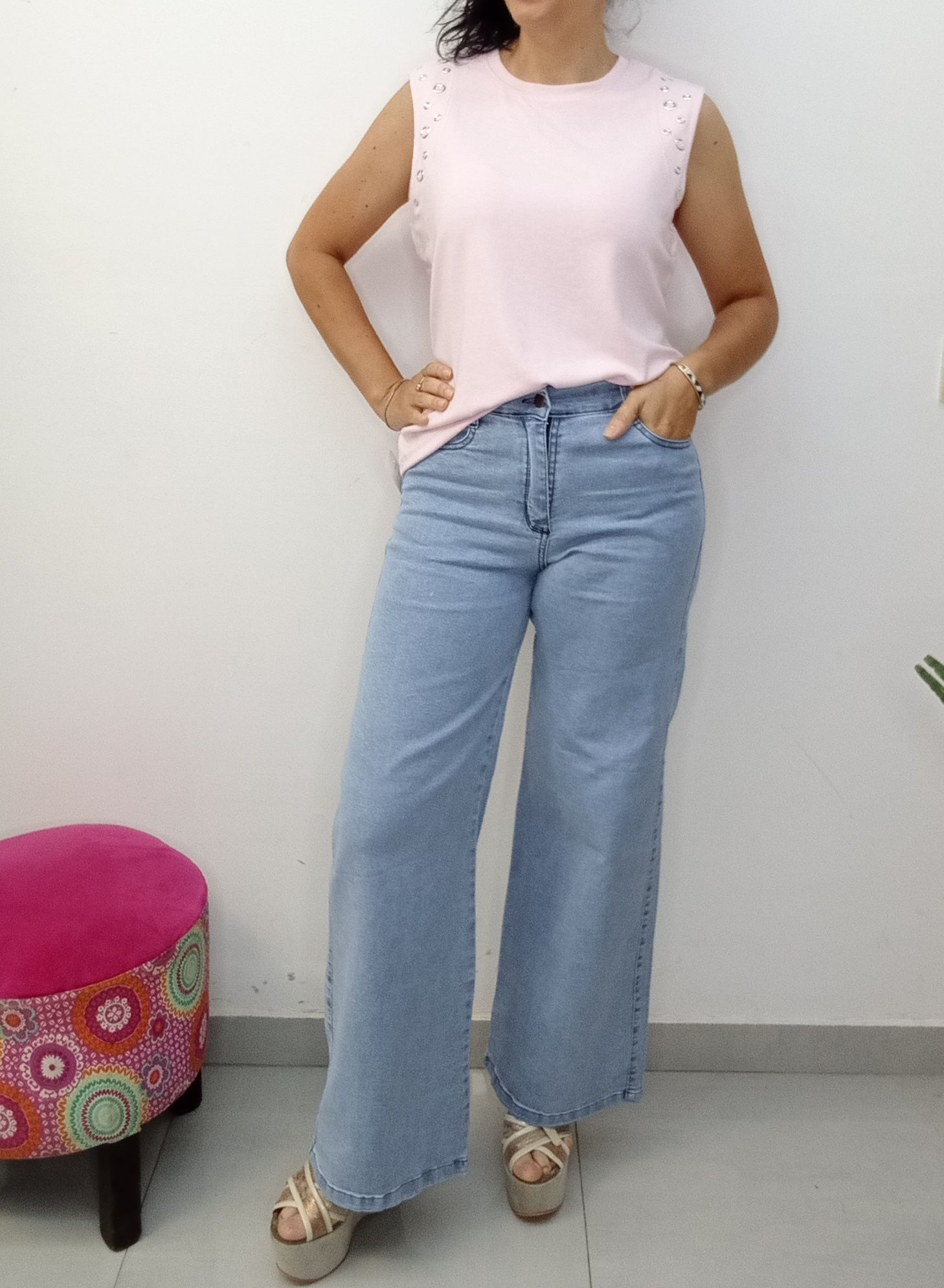 Mujer con top rosa y vaqueros anchos, con las manos en las caderas. Sandalias de plataforma beige, pared blanca.