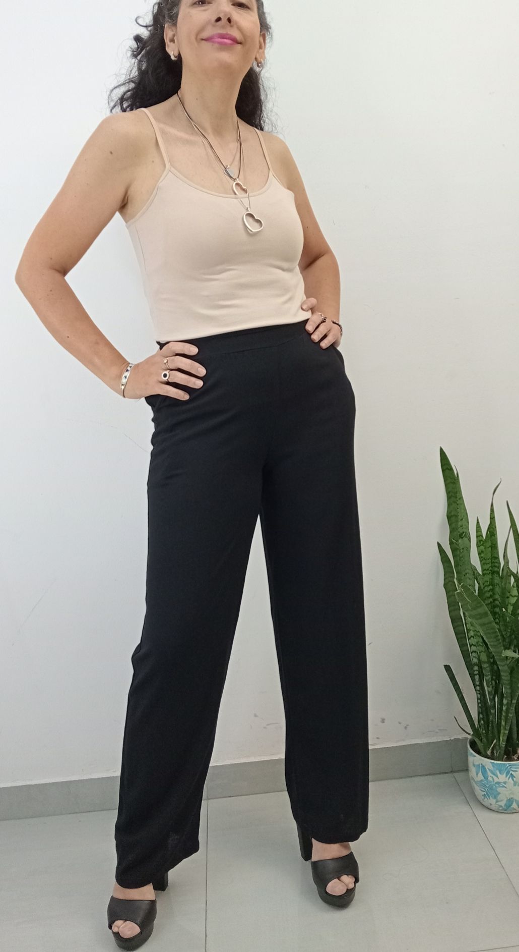 Mujer vestida con pantalones negros y una camisola color canela, sonriendo con las manos en las caderas.