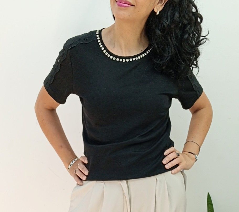 Mujer con camisa negra con escote joya y pantalones color canela, manos en las caderas, sonriendo.