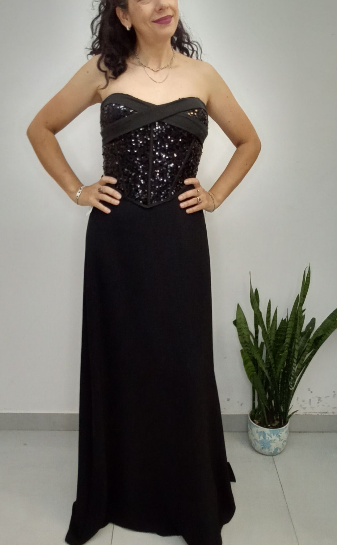 Mujer con un vestido negro sin tirantes con lentejuelas, con las manos en las caderas, sonríe; entorno interior.