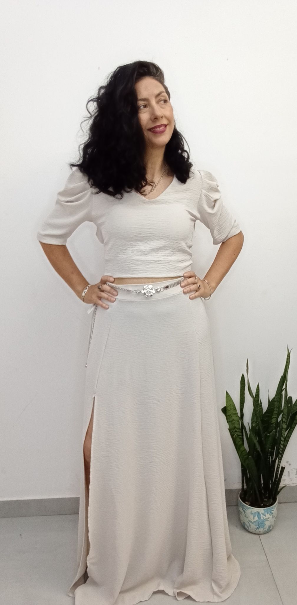 Mujer con un traje de dos piezas de color crema y falda con abertura alta, posando frente a una pared blanca.