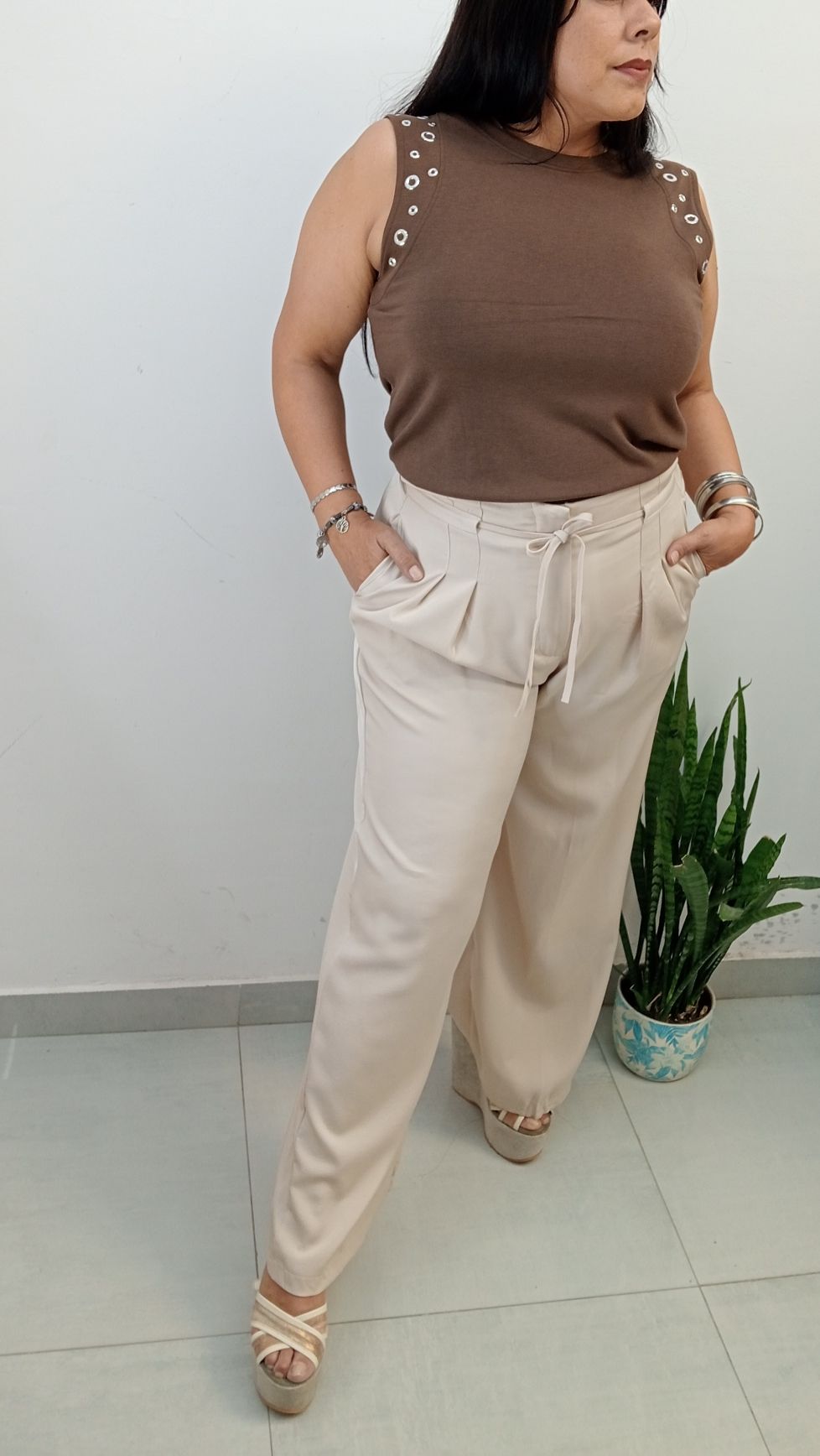 Mujer con top marrón y pantalones color canela, con las manos en los bolsillos, posando junto a una planta.