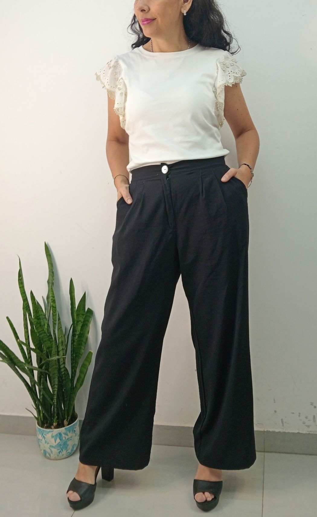 Mujer con top blanco y pantalones negros de pierna ancha con las manos en los bolsillos, de pie junto a una planta.