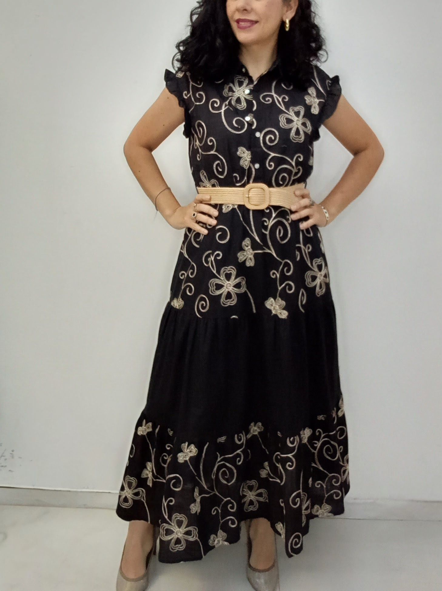 Mujer con un vestido negro con bordados florales, ceñido con un cinturón beige, de pie en el interior.