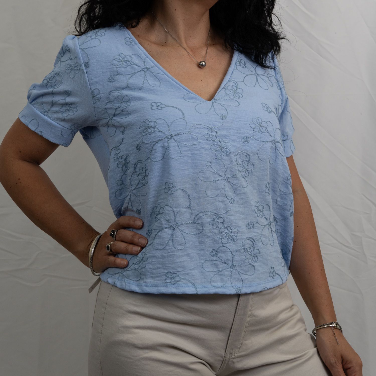Mujer con una camisa azul de cuello en V con bordados y pantalones beige, posando con la mano en la cadera.