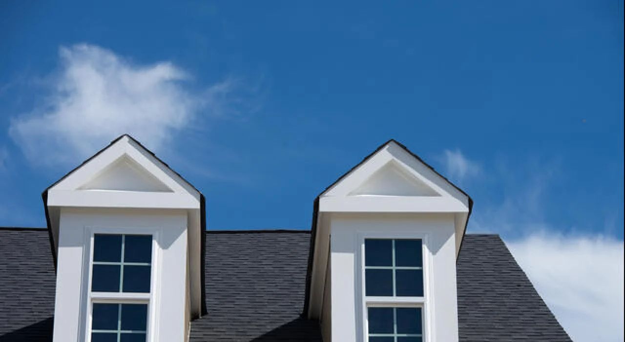 Two white dormer windows on a dark roof beneath a blue sky with clouds