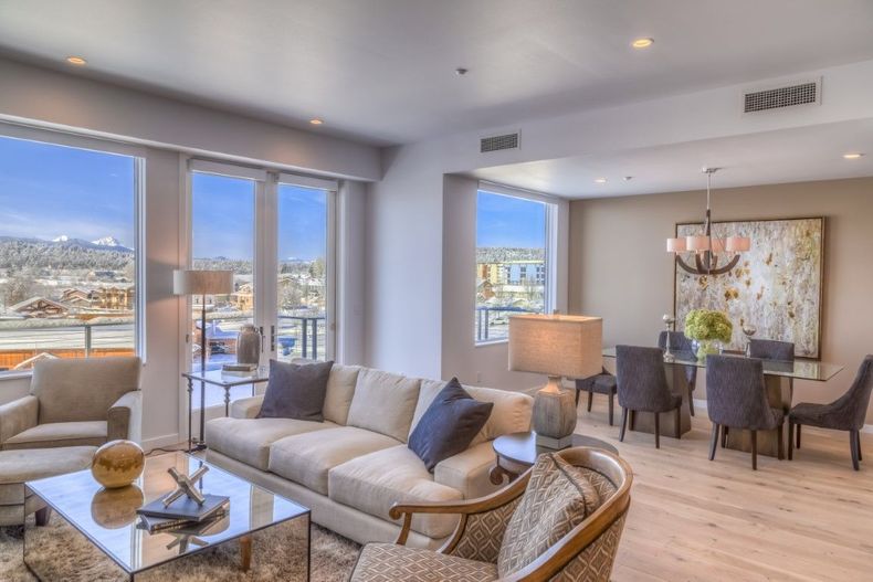 Bright modern living room with beige sofas, dining area, and large windows overlooking a city view