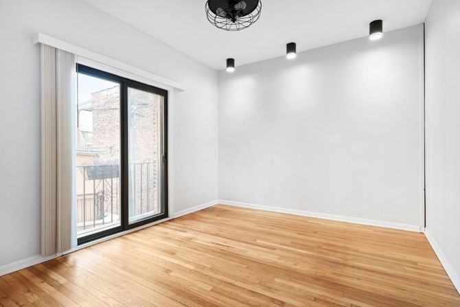 Empty modern room with hardwood floors, white walls, ceiling lights, and a sliding glass door to a balcony