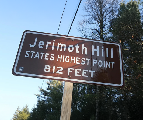 Brown roadside sign reading “Jerimoth Hill, States Highest Point, 812 Feet” against trees and blue sky