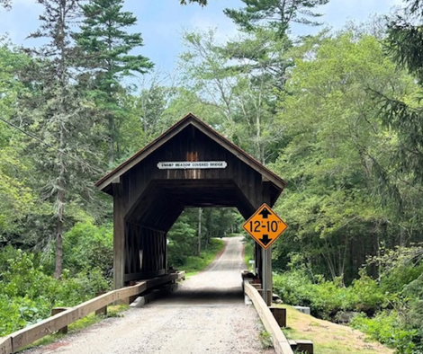 Covered wooden bridge over a gravel road in a forest, with a 12'10